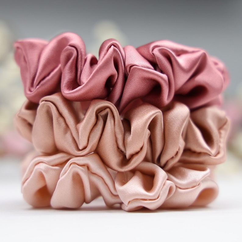 May include: Three stacked hair scrunchies in shades of pink and peach. The top scrunchie is a deep rose color, the middle scrunchie is a light peach color, and the bottom scrunchie is a pale pink color.