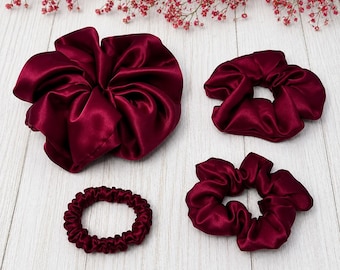 May include: A set of four deep burgundy hair accessories made of a smooth, satin-like fabric. The collection includes one extra-large ruffled scrunchie, two standard-sized ruffled scrunchies, and one thin, gathered hair tie, all in a matching dark red.