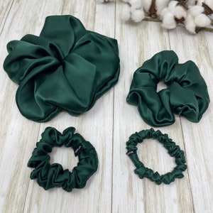 May include: Three green satin scrunchies of different sizes. The largest scrunchie is a large, loose bun style. The medium scrunchie is a classic, gathered style. The smallest scrunchie is a thin, gathered style. All scrunchies have a black tag that reads "100% silk".