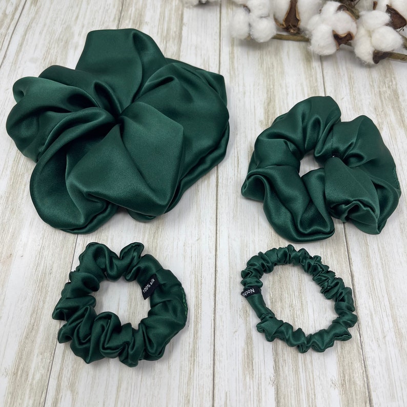 May include: Three green satin scrunchies of different sizes. The largest scrunchie is a large, loose bun style. The medium scrunchie is a classic, gathered style. The smallest scrunchie is a thin, gathered style. All scrunchies have a black tag that reads "100% silk".