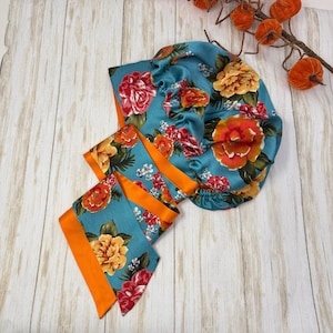 Op de afbeelding: Een blauwe satijnen haarbonnet met een bloemenprint in tinten oranje, rood en geel. De bonnet is vastgebonden met een bijpassend oranje satijnen lint.