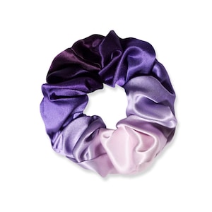 May include: A circular hair scrunchie made of smooth, shiny fabric featuring a gradient color transition. The colors shift from a deep, dark purple at the top to a lighter lavender and finally a pale, soft lilac shade at the bottom edge.