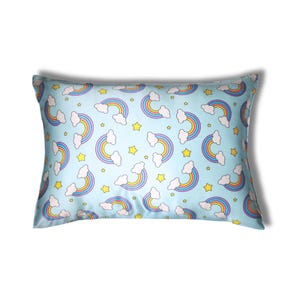 May include: A rectangular pillow with a light blue background featuring a repeating pattern of rainbows, white clouds, and yellow stars. The rainbows have multiple colors, and the pillow has a satin-like sheen.