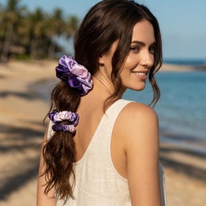 May include: A set of three hair scrunchies in varying shades of purple and lavender. The largest scrunchie features a satin-like finish with a gathered texture, while the two smaller scrunchies display a matching gradient of light to dark purple.