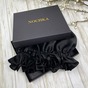 May include: A black satin hair bonnet with ruffles, packaged in a black box with the word "NOCHKA" printed in gold lettering.