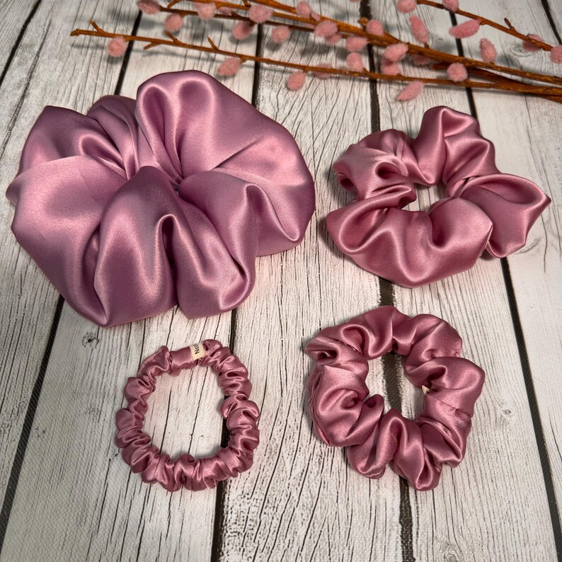May include: Four pink satin scrunchies in different sizes. The largest scrunchie is a large, loose bun style. The other three are smaller and more compact.