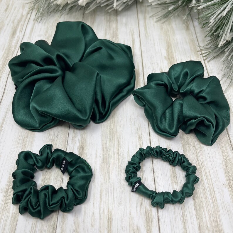 May include: Four green satin scrunchies of different sizes. The largest scrunchie is in the center of the image, and the smallest scrunchie is in the lower left corner. The scrunchies are all made of a soft, silky fabric.
