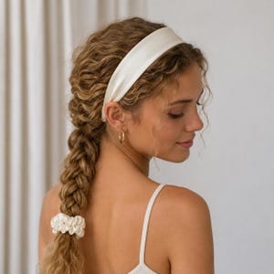 May include: A smooth, off-white fabric headband worn across the top of the head, paired with a matching off-white ruffled fabric scrunchie securing the end of a long, thick, braided hairstyle. The accessories feature a soft, satin-like sheen.