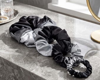 May include: A set of three hair scrunchies in varying sizes, each featuring a smooth, lustrous fabric with a gradient color transition from black to silver-gray. The smallest scrunchie includes a small black fabric tag labeled with the text Nochka.