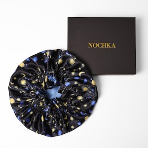 May include: Black satin bonnet with a celestial pattern of gold suns, moons, and stars, and blue accents. The bonnet is next to a black box with the gold text "NOCHKA".