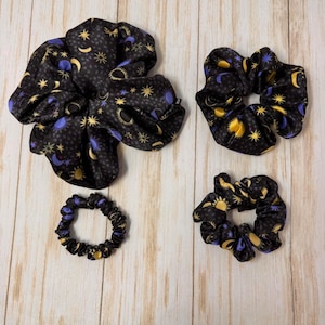 May include: Three black satin scrunchies with a celestial print featuring moons, stars, and suns in shades of yellow and purple.