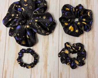 May include: Three black satin scrunchies with a celestial print featuring moons, stars, and suns in shades of yellow and purple.