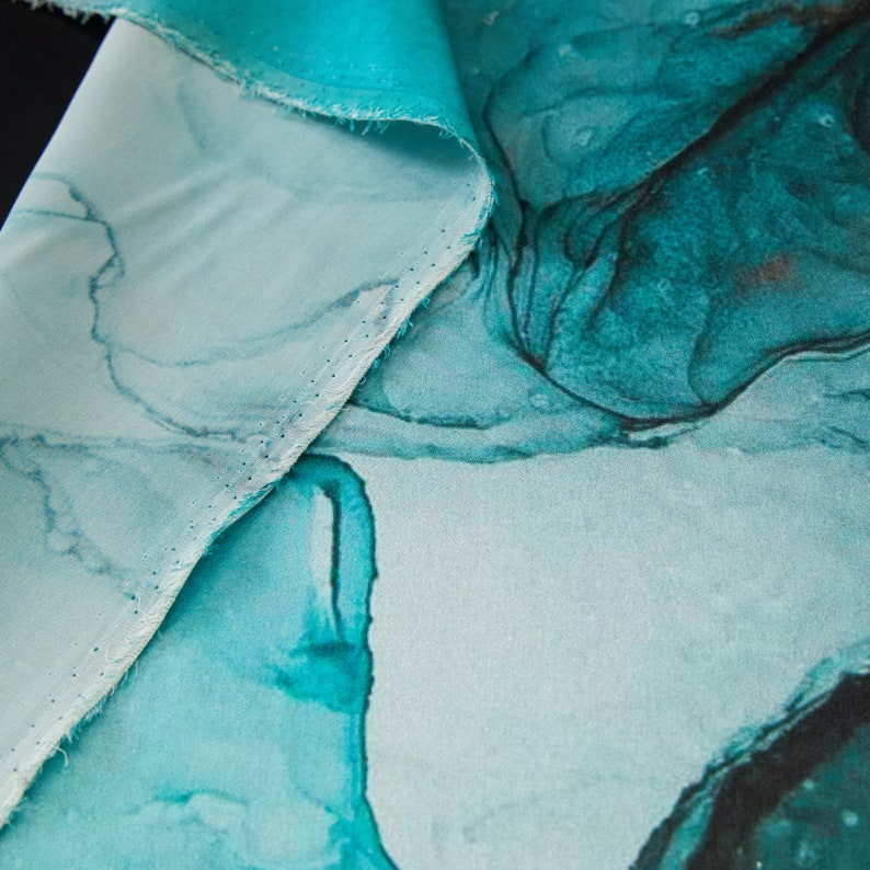 May include: Close-up of a teal blue fabric with a white background and a marbled pattern. The fabric has a subtle sheen and a soft texture.
