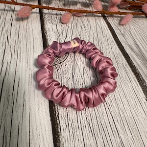 May include: A dusty rose colored scrunchie made of silky fabric. The scrunchie is in a circle shape and has a small tag with the word "MIN" on it.