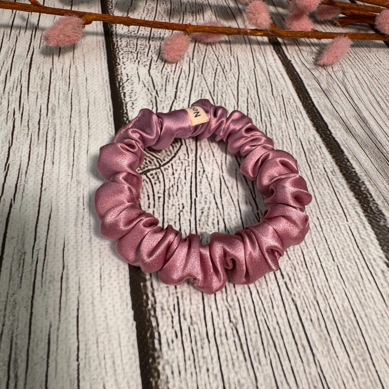 May include: A dusty rose colored scrunchie made of silky fabric. The scrunchie is in a circle shape and has a small tag with the word "MIN" on it.