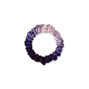 May include: A circular hair scrunchie featuring a smooth, satin-like fabric with a gradient color transition. The color palette shifts from a deep, dark purple at the bottom to a light, soft lavender and pale pink at the top, creating a continuous ombre effect.