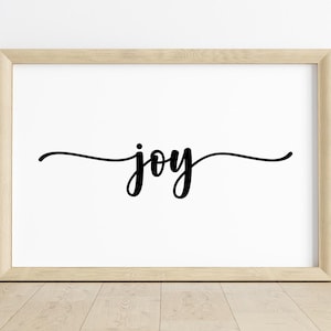 Puede incluir: Una impresión enmarcada con la palabra "joy" en escritura cursiva negra centrada en un fondo blanco. El marco es de color madera clara, y la impresión se muestra sobre un suelo de madera.