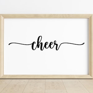 May include: A framed print with the word "cheer" in elegant black script. The word is centered on a white background, set within a light-colored wooden frame. The print is displayed on a wooden floor.