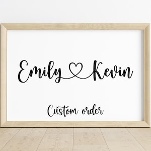 Pode incluir: Uma impressão emoldurada com os nomes "Emily" e "Kevin" em escrita preta, conectados por um símbolo de coração. Abaixo dos nomes, as palavras "Custom order" estão escritas em uma fonte cursiva menor. A moldura é de cor de madeira clara.