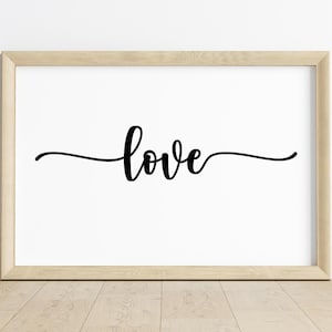 Puede incluir: Una impresión enmarcada con la palabra "love" en escritura cursiva negra centrada en un fondo blanco. El marco es de madera clara y la impresión se muestra sobre un suelo de madera.