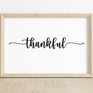 Pode incluir: Uma impressão emoldurada com a palavra "thankful" em elegante escrita preta sobre um fundo branco. A moldura é de cor de madeira clara, e a impressão é exibida em um piso de madeira contra uma parede branca.