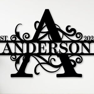 Op de afbeelding: Zwart metalen familienaambord met de naam "Anderson" en de letter "A" als middelpunt. Het bord bevat de tekst "Est. 2025" en decoratieve krullen. De woorden "Custom Order" zijn ook zichtbaar.