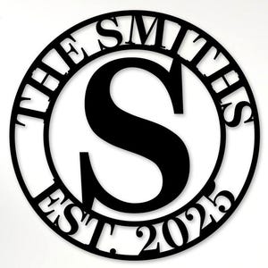 May include: Circular black metal sign featuring the name "The Smiths", the letter "S", and the year "EST 2025". The sign is mounted on a white wall. The words "Custom Order" are also visible.