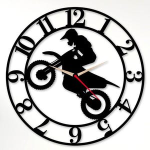 Peut inclure: Horloge murale en métal noir représentant une silhouette de pilote de motocross sur une moto. L'horloge a un design circulaire avec des chiffres et des aiguilles noirs, et une trotteuse rouge. L'horloge est fixée sur un mur blanc.