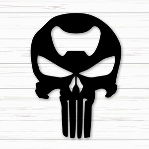 May include: Black bottle opener in the shape of a skull with a bottle cap on the bottom. The skull design has a large opening for the bottle cap and a detailed facial structure. The background is a white wooden surface.