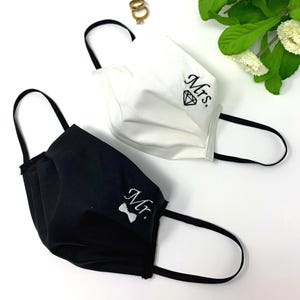 Puede incluir: Dos mascarillas, una negra y otra blanca, con presillas negras. La mascarilla negra tiene bordado "Mr." con una pajarita, y la mascarilla blanca tiene "Mrs." y un diamante. Anillos dorados y flores blancas en el fondo.
