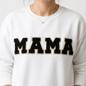 May include: White sweatshirt with the word "MAMA" in large, black, textured letters with gold trim across the chest. The sweatshirt has long sleeves and a crew neck. The person wearing the sweatshirt is smiling.