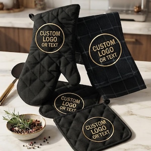 May include: A set of black kitchen textiles including a quilted oven mitt, a square pot holder, and a rectangular towel, each featuring a circular gold emblem with the text CUSTOM LOGO OR TEXT. The items are arranged on a white marble surface.