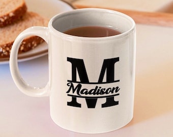 May include: White ceramic mug with a black letter "M" and the name "Madison" printed on it. The mug is filled with a brown liquid.