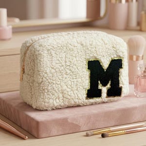May include: A cream-coloured, fuzzy sherpa cosmetic bag featuring a large, black, varsity-style letter M patch with a gold glitter border on the front. The rectangular pouch has a gold-toned zipper closure along the top edge.