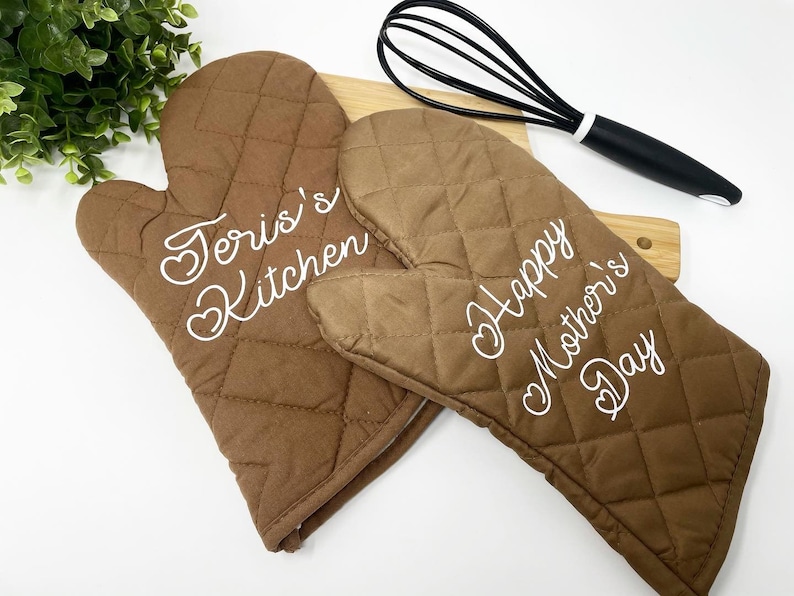 May include: Two brown quilted oven mitts with white embroidered text. One mitt says "Teri's Kitchen" and the other says "Happy Mother's Day".