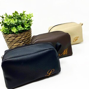 May include: Three zippered travel pouches in black, brown, and beige with a textured leather-like finish. Each pouch has a single initial embroidered in orange on the front: L, M, and F.