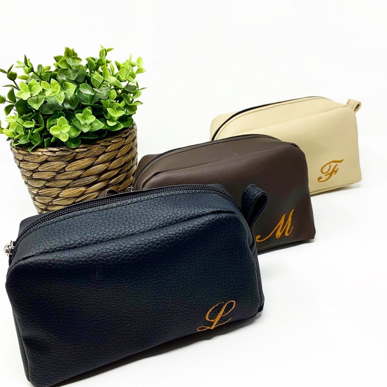 May include: Three zippered travel pouches in black, brown, and beige with a textured leather-like finish. Each pouch has a single initial embroidered in orange on the front: L, M, and F.