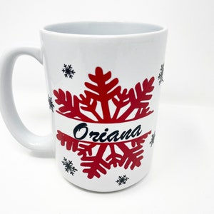 May include: White ceramic mug with a red snowflake design and the name "Oriana" written in black.