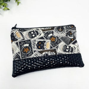 Puede incluir: Bolsa de cremallera negra y blanca con un estampado negro y naranja de cartas de tarot, calaveras y gatos. La bolsa tiene un fondo negro texturizado.