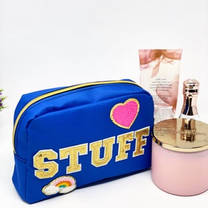 Op de afbeelding: Een blauwe toilettas met rits, een gouden glitterpatch met de tekst "STUFF", een roze hartpatch en een regenboogpatch. De tas ligt op een wit oppervlak naast een roze kaars en een fles lotion.
