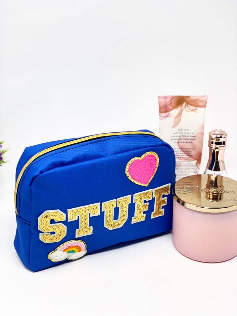 Op de afbeelding: Een blauwe toilettas met rits, een gouden glitterpatch met de tekst "STUFF", een roze hartpatch en een regenboogpatch. De tas ligt op een wit oppervlak naast een roze kaars en een fles lotion.