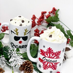 May include: Two white ceramic mugs with festive designs. One mug features a reindeer with sunglasses and the name "Ricardo" and the other mug has a red snowflake with the name "Oriana". Both mugs are filled with hot chocolate topped with whipped cream and chocolate chips.