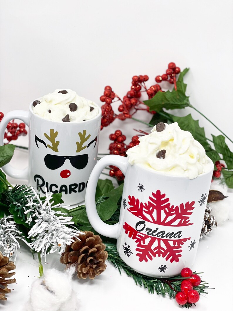 May include: Two white ceramic mugs with festive designs. One mug features a reindeer with sunglasses and the name "Ricardo" and the other mug has a red snowflake with the name "Oriana". Both mugs are filled with hot chocolate topped with whipped cream and chocolate chips.