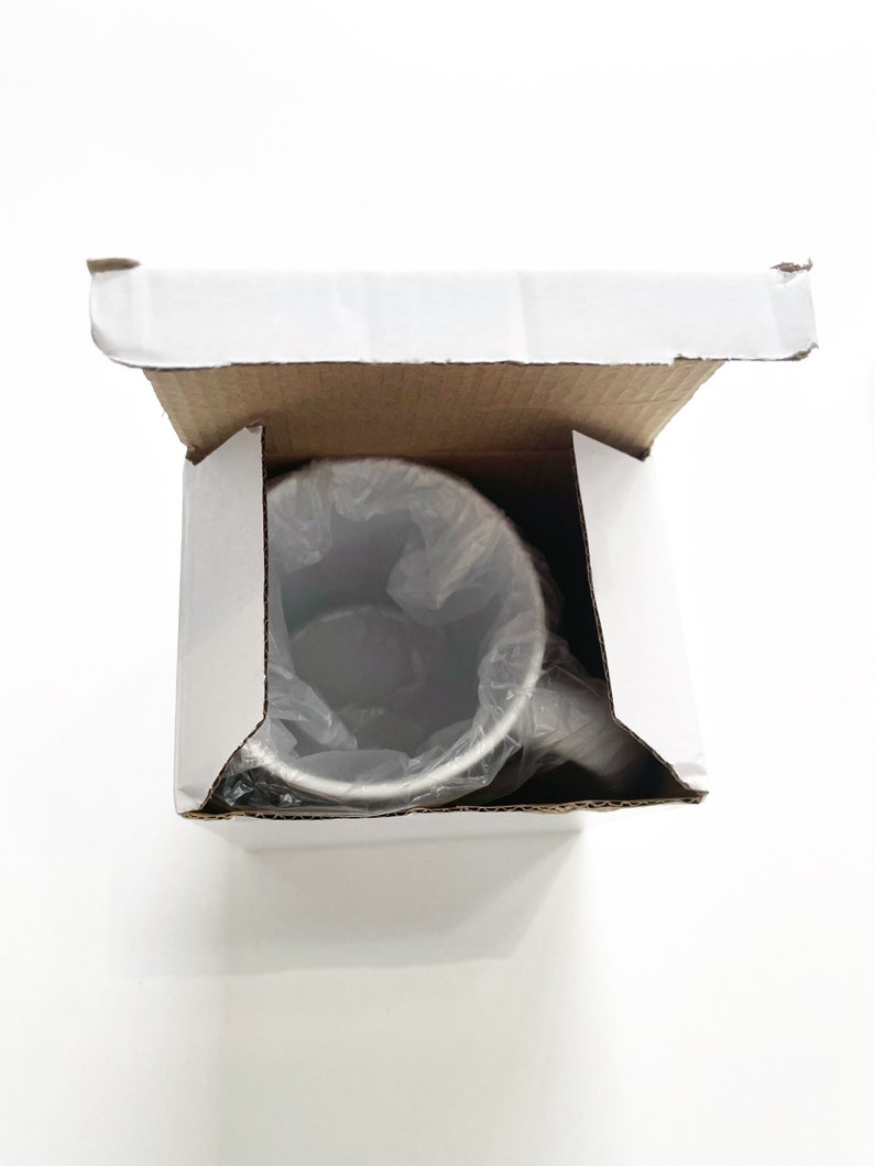 May include: A white cardboard box with the top open, revealing a silver stainless steel travel mug inside. The mug is wrapped in clear plastic.