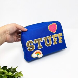 Op de afbeelding: Een koninklijk blauwe ritszak met het woord "STUFF" in gouden glitterletters. De tas heeft een roze hart en een regenboogpatch.