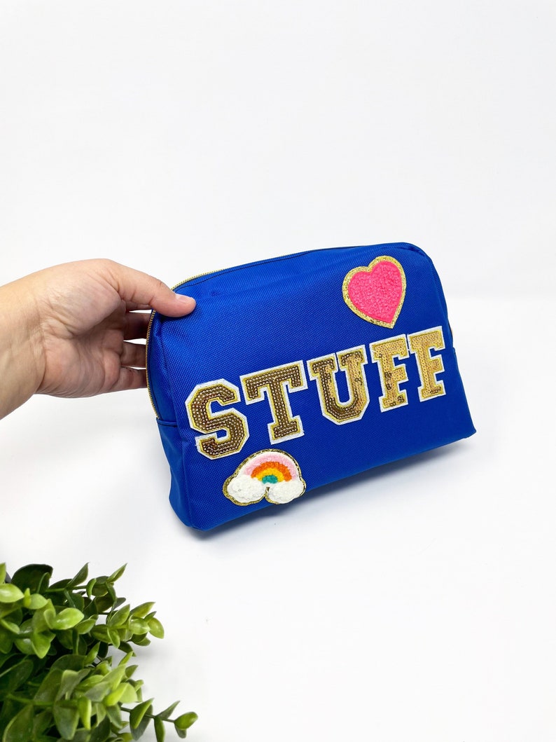 Op de afbeelding: Een koninklijk blauwe ritszak met het woord "STUFF" in gouden glitterletters. De tas heeft een roze hart en een regenboogpatch.