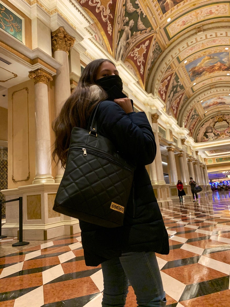 May include: A person wearing a black jacket, blue jeans, and a black face mask is holding a black quilted tote bag with a gold logo that says "OMMO".