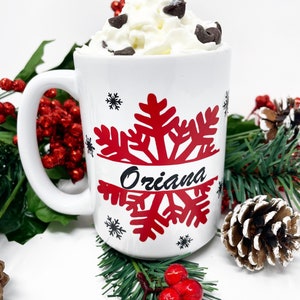 May include: White ceramic mug with a red snowflake design and the name "Oriana" written in black. The mug is filled with a white beverage topped with whipped cream and chocolate chips. The mug is surrounded by green pine branches and red berries.