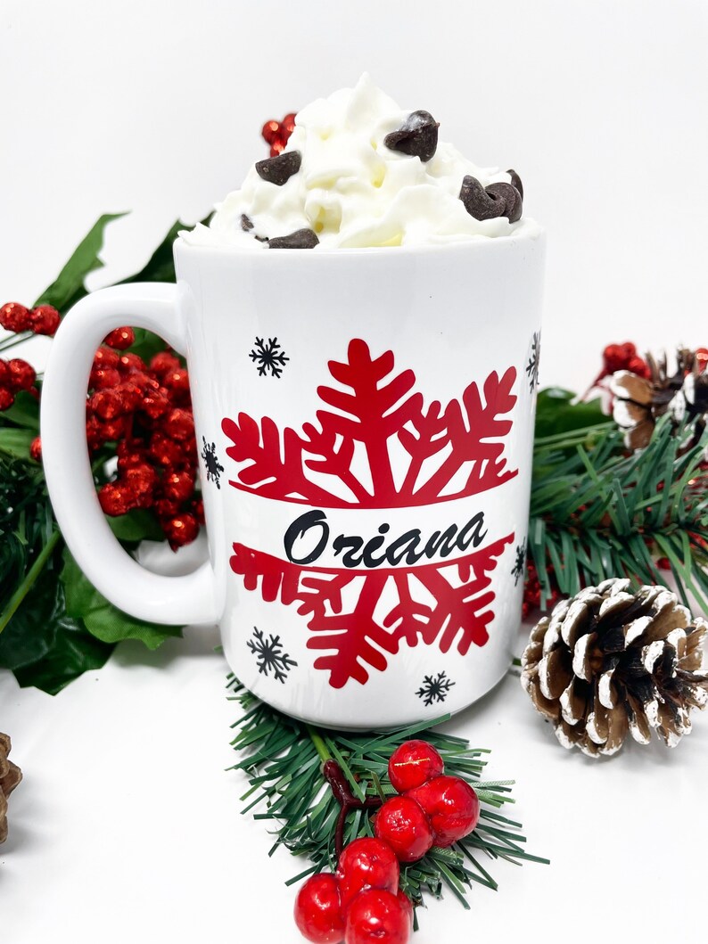 May include: White ceramic mug with a red snowflake design and the name "Oriana" written in black. The mug is filled with a white beverage topped with whipped cream and chocolate chips. The mug is surrounded by green pine branches and red berries.