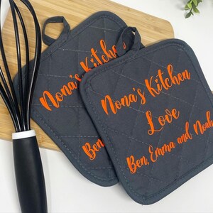 May include: Two gray quilted pot holders with orange text. The text on one pot holder reads "Nona's Kitchen Love Ben, Emma and Noah". The text on the other pot holder reads "Nona's Kitchen".